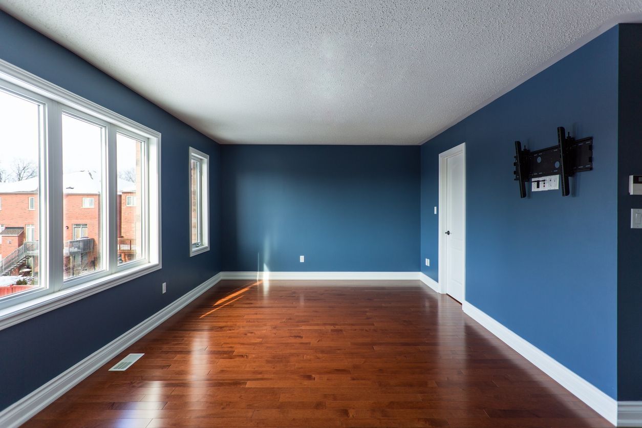 Empty room with blue walls, hardwood floor, and large windows on the left.