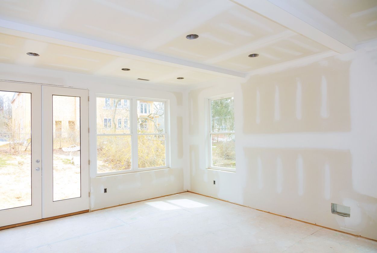 Bright empty room with white walls, large windows, and unfinished drywall patches