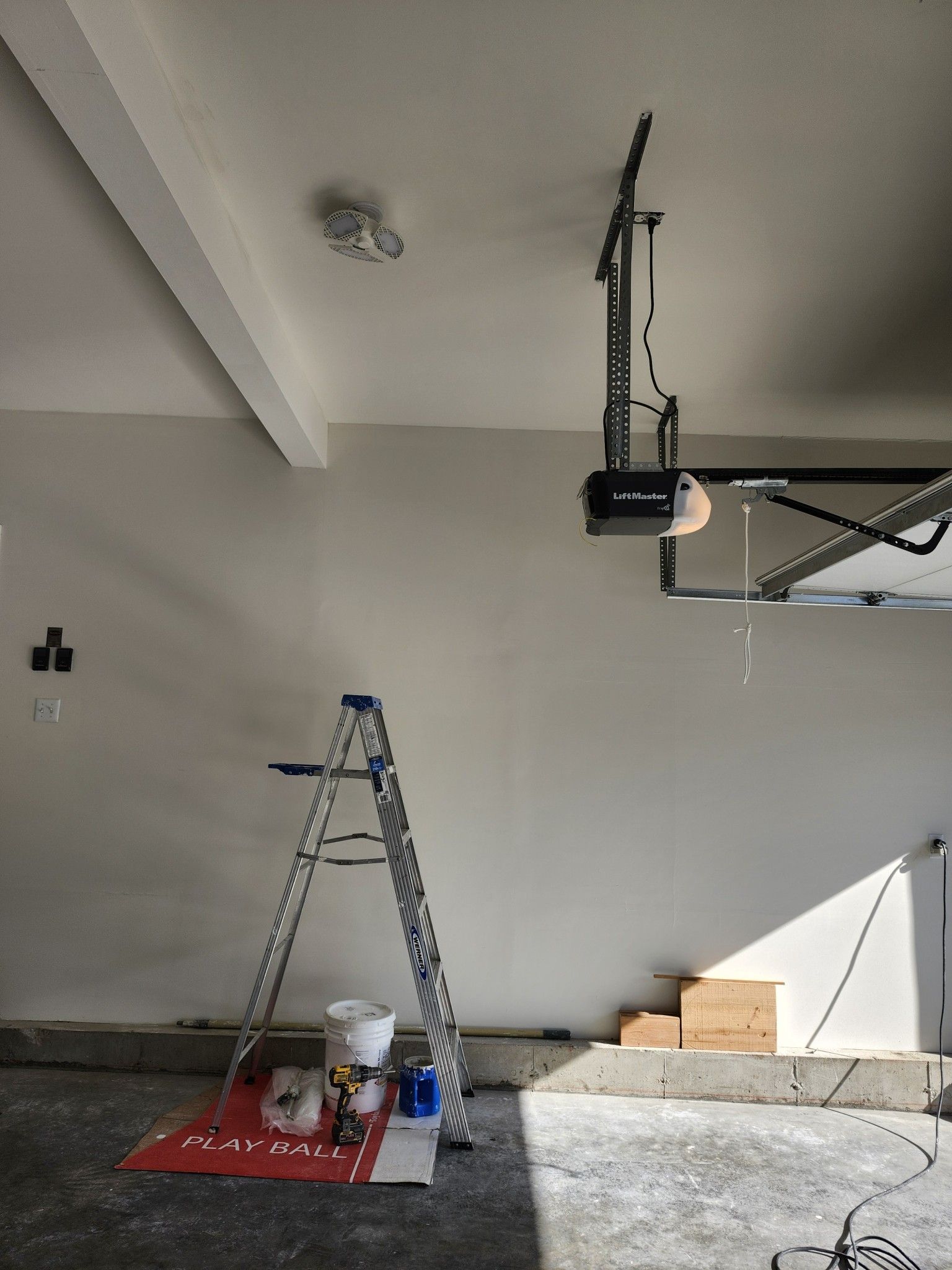 Ladder and paint supplies in a garage under renovation, with a garage door opener and unfinished steps.