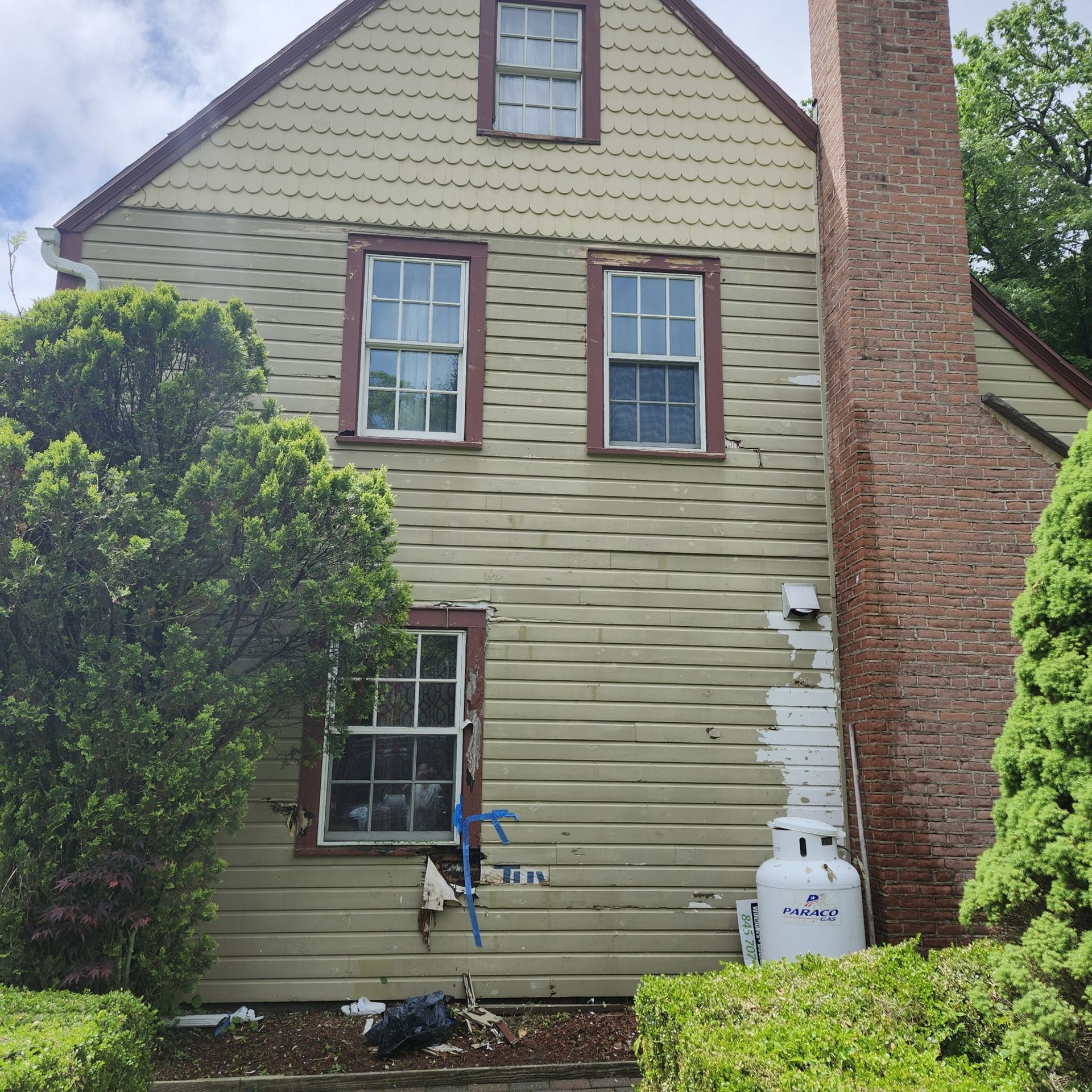 Rear view of a tan house with two windows, a brick chimney, and a white propane tank beside the wall.