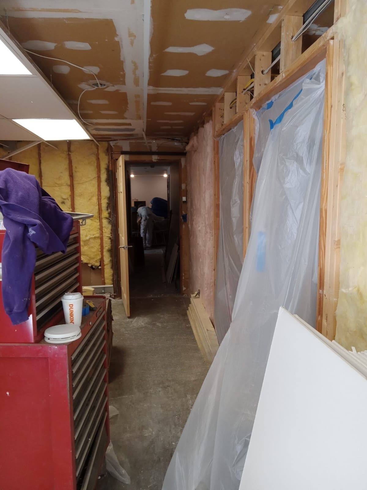 Narrow unfinished hallway with exposed drywall, plastic sheeting, and renovation materials along the walls.