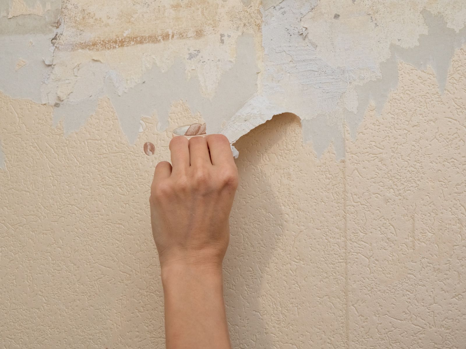 Hand scraping peeling paint from a beige wall with a small tool
