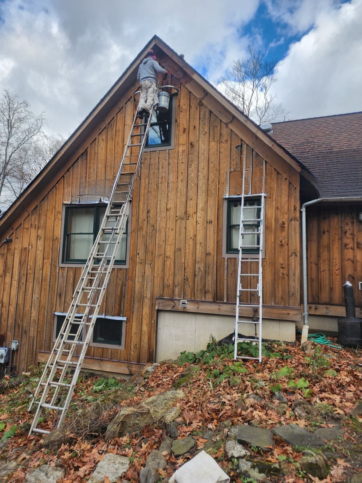 Ladder leaning to attic window on wooden house; another ladder stands below, with fall leaves and cloudy sky.