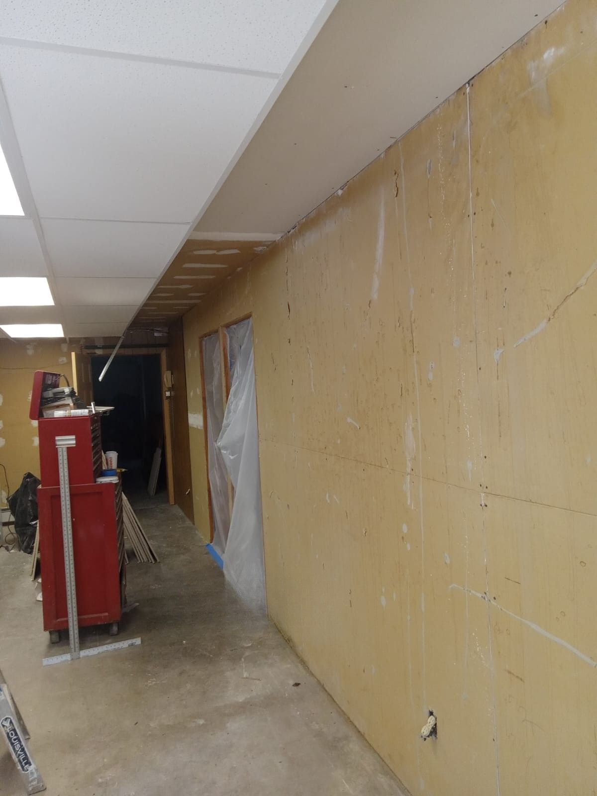 Unfinished hallway with drywall on the right, ceiling tiles above, and construction equipment on the left.