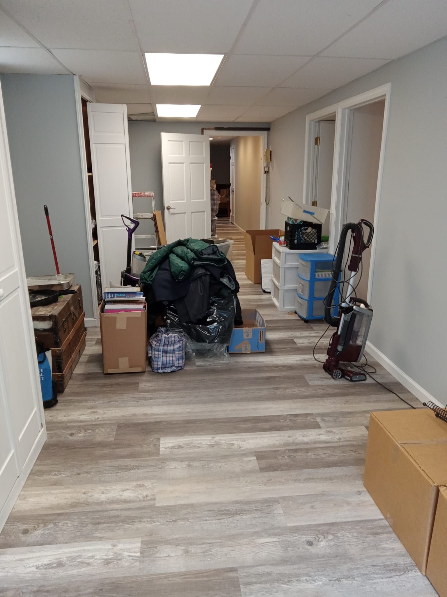 Cluttered hallway with boxes, bags, and cleaning supplies in an indoor office or storage area.