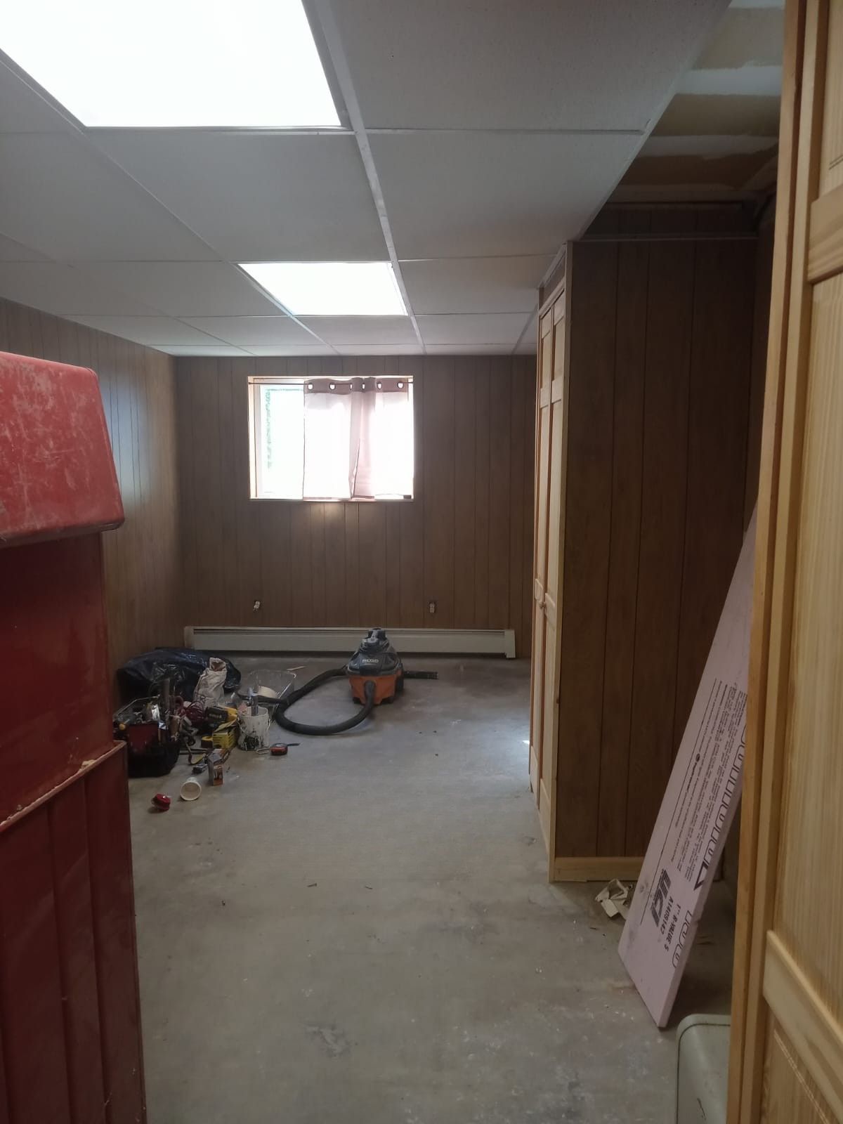 Small unfinished room with wood-paneled walls, ceiling tiles, window, and scattered tools on the floor.