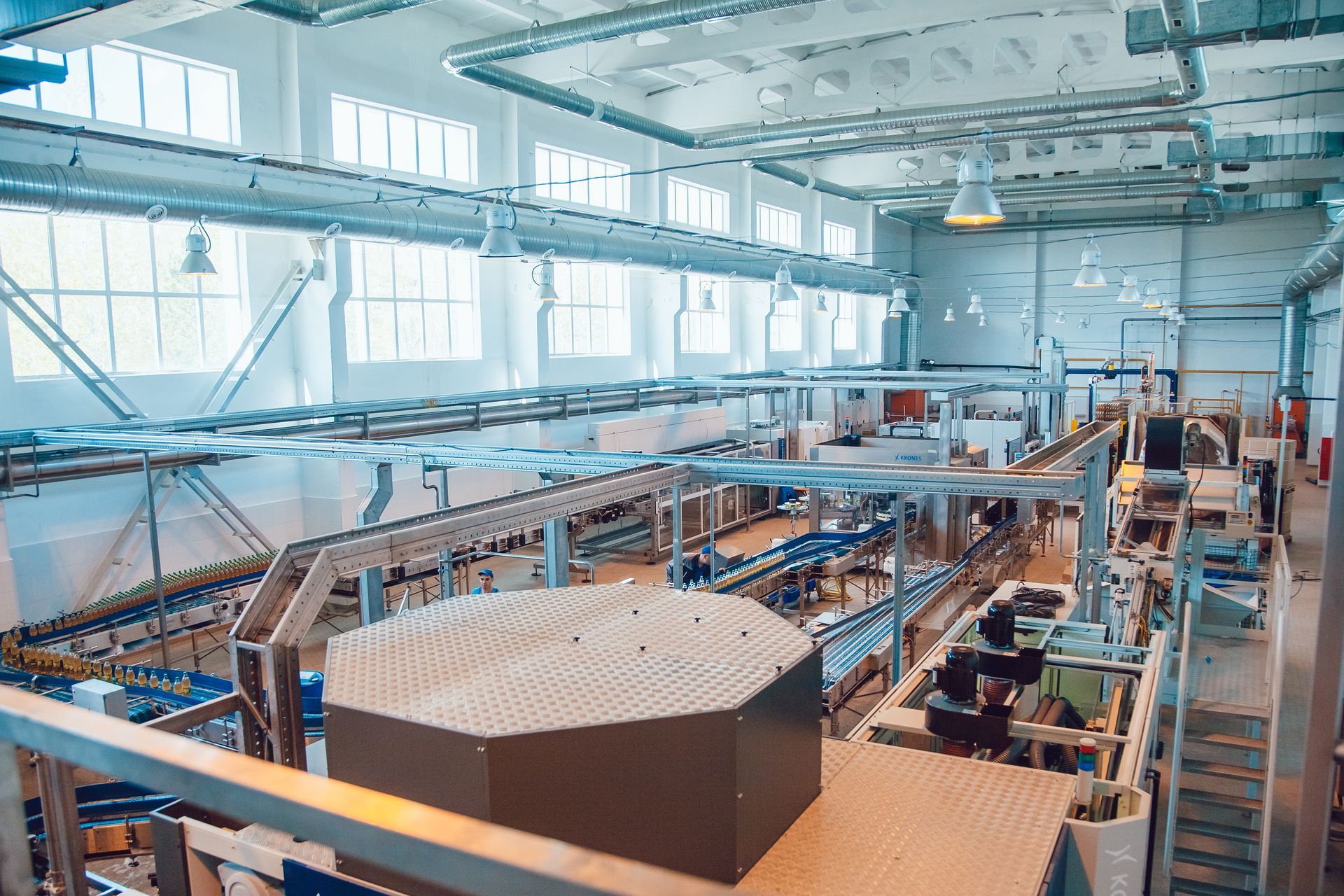 Person steam cleaning white tiled floor with steam cleaner, wearing gloves. Factory interior with conveyor belts, overhead lighting, and large container.