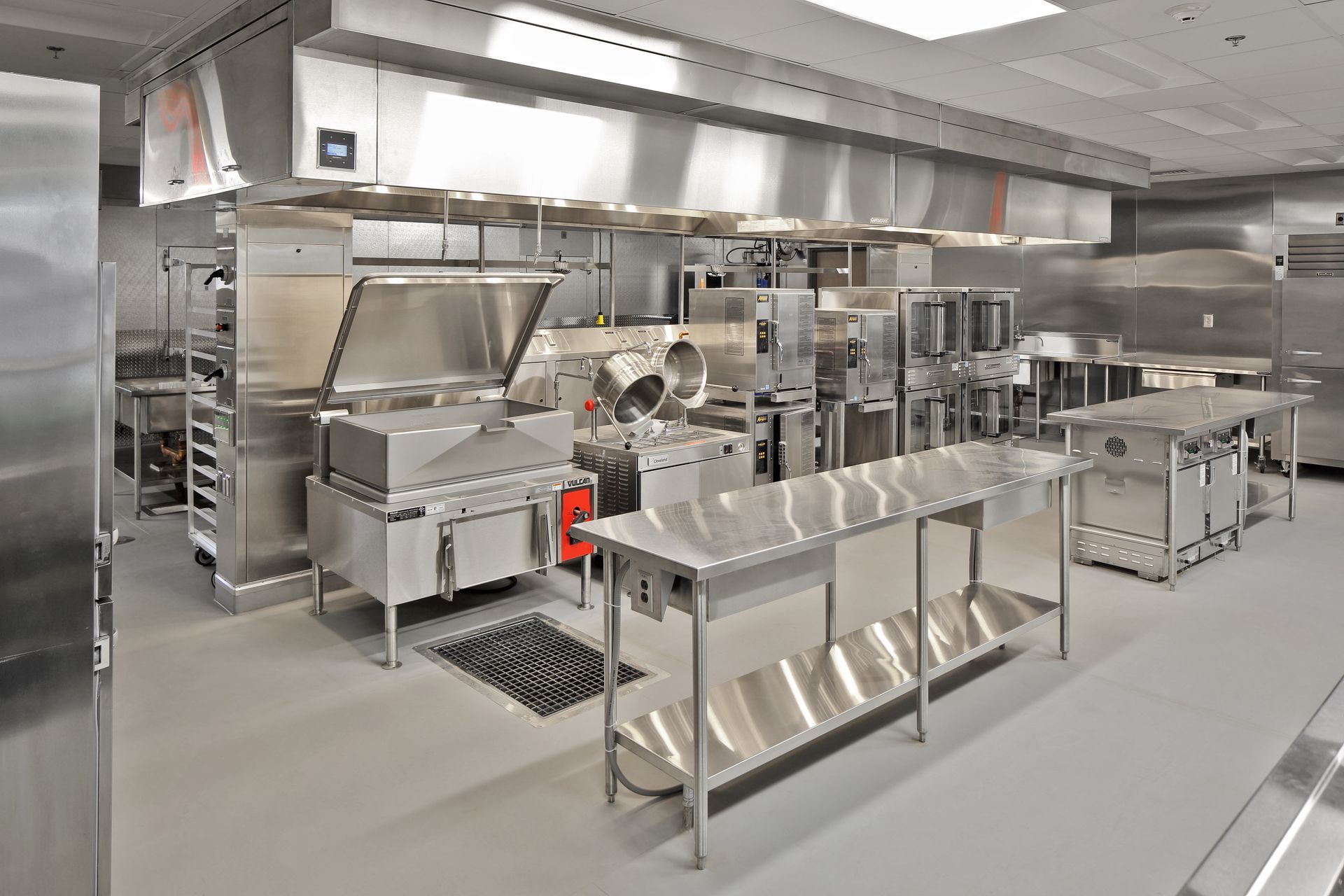 Person in yellow gloves cleaning oven with a yellow steam cleaner. A commercial kitchen with stainless steel equipment and a gray floor.