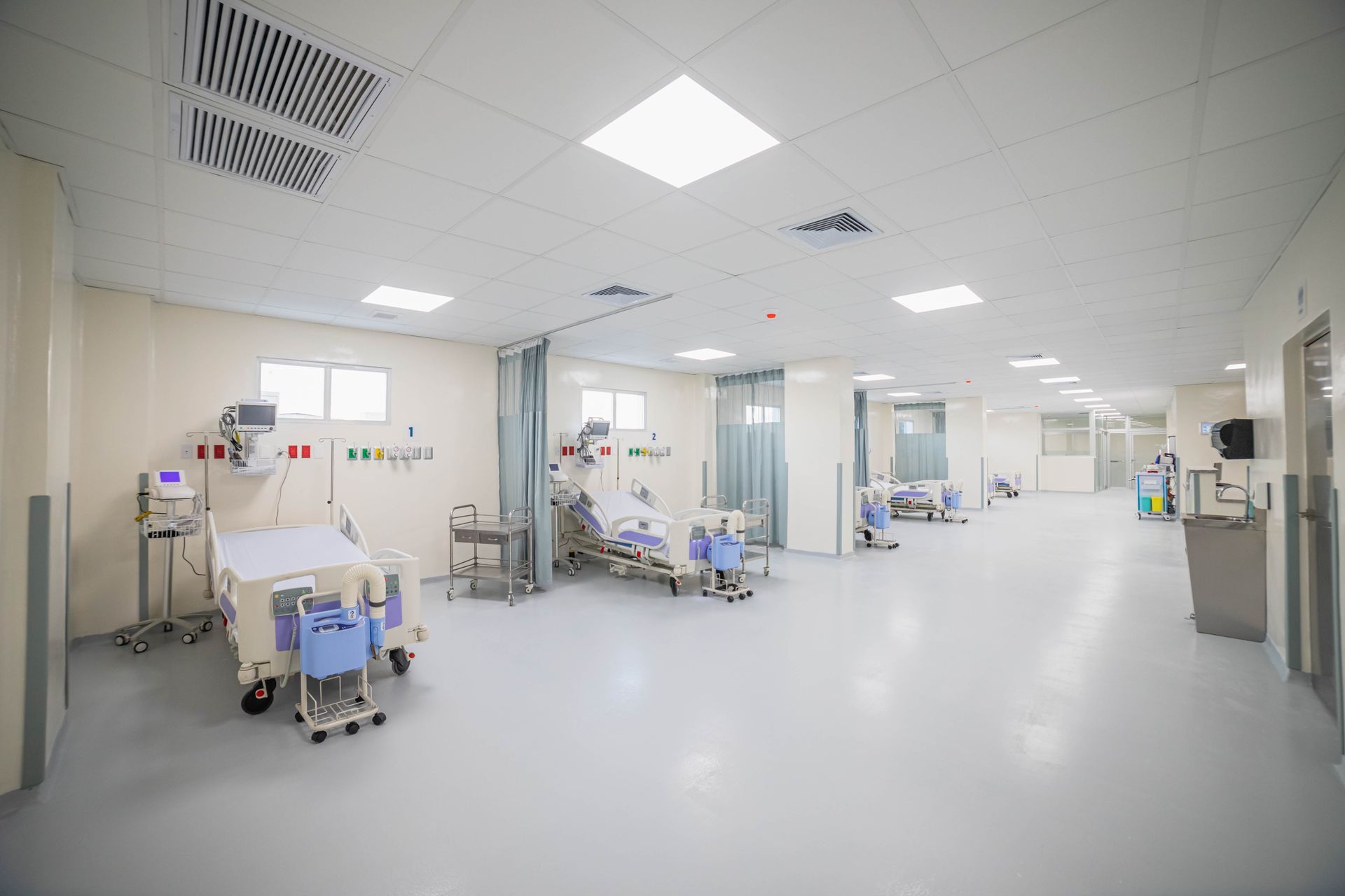 Hand using a vacuum cleaner head to clean a teal couch. Hospital room with beds, medical equipment, and a long hallway.