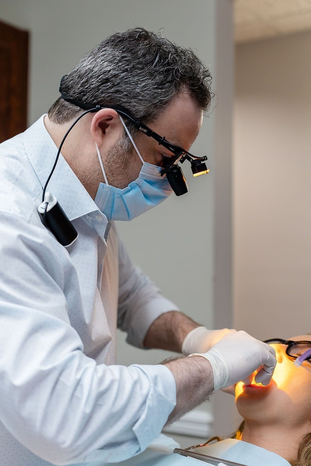Dentist in white coat and mask examining a patient with a dental light