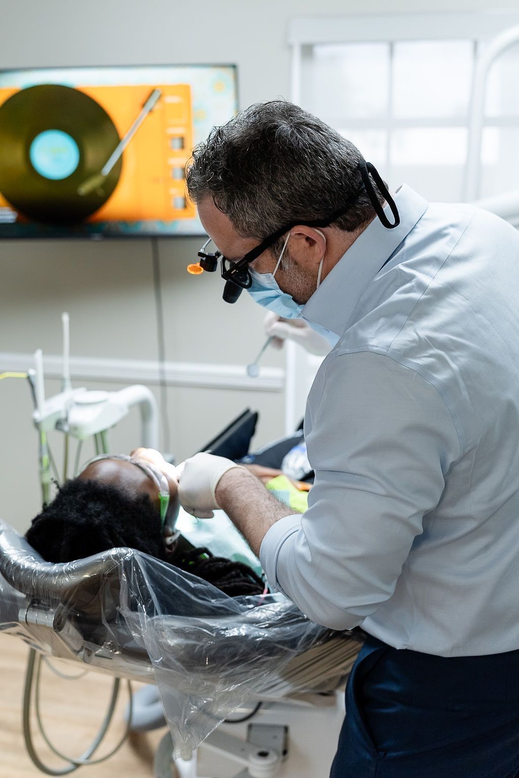 Dentist wearing magnifying glasses examining a patient in a dental chair, with a monitor in the background