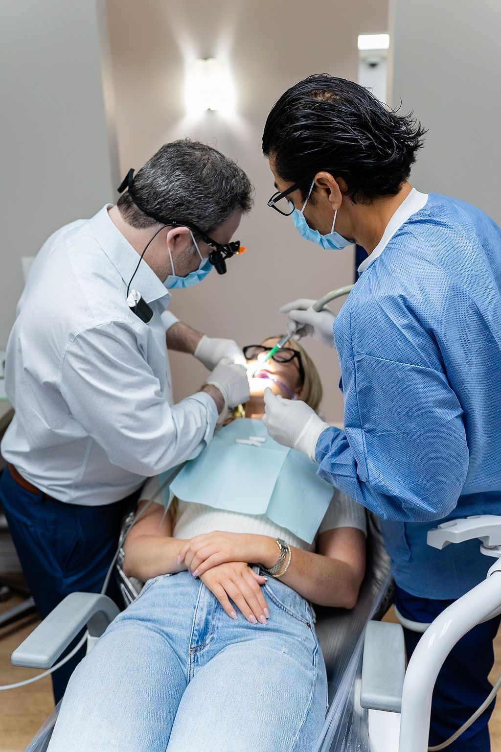 Two dentists treating a patient in a dental chair, wearing masks and gloves, in a clinic room.