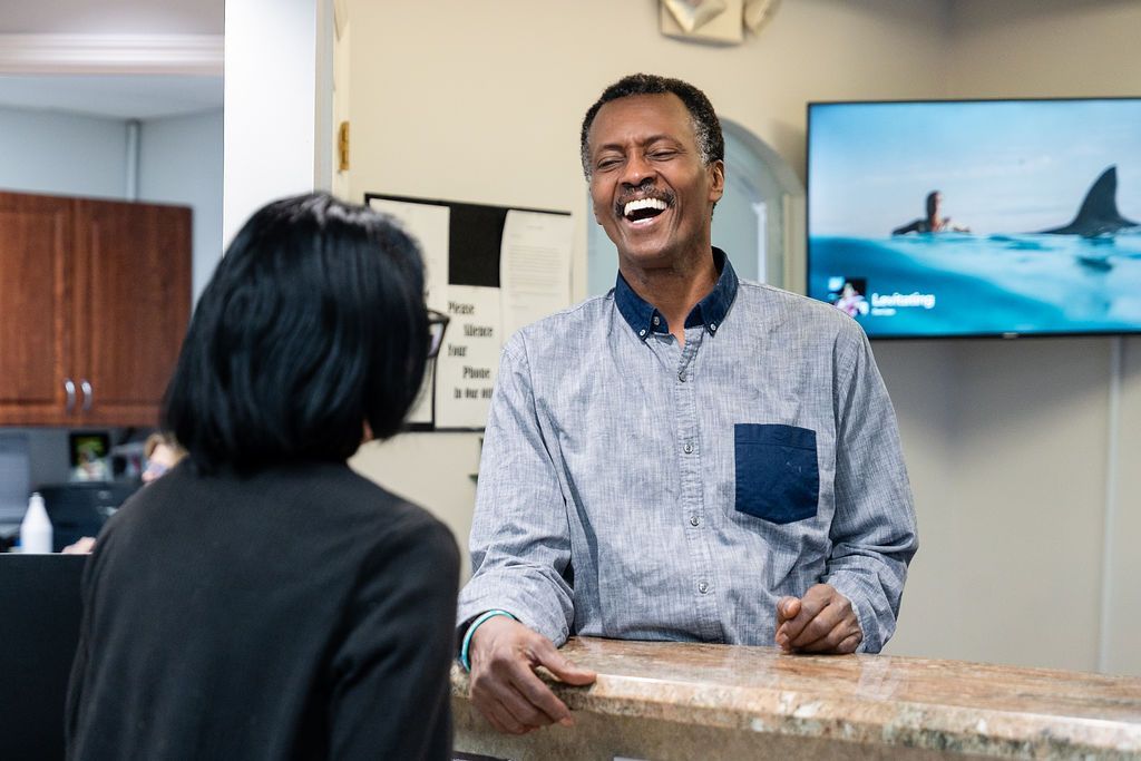 Man smiling behind a reception counter, speaking with a woman in an office lobby.