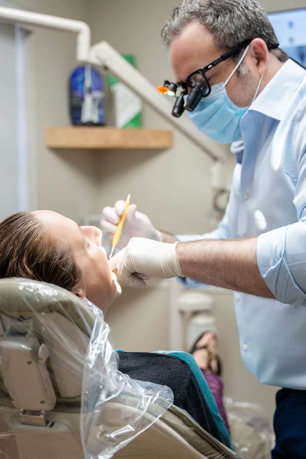 Dentist treating a patient in a chair, wearing gloves, mask, and magnifying glasses