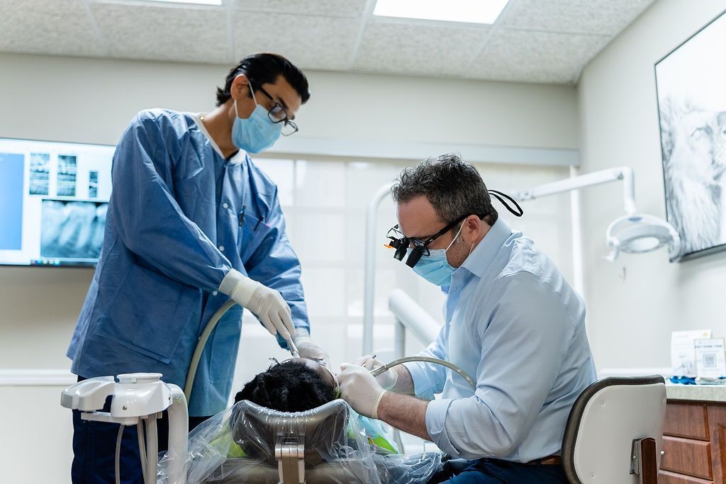 Two dental clinicians in masks and gloves treat a patient in a bright exam room.