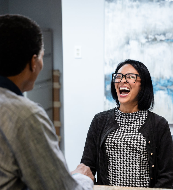 Two people smiling and shaking hands in an office, with one laughing and wearing glasses.