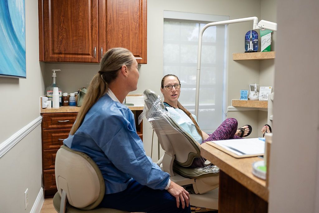 Two people sitting in a dental exam room, one reclined in a chair near a desk and cabinets.