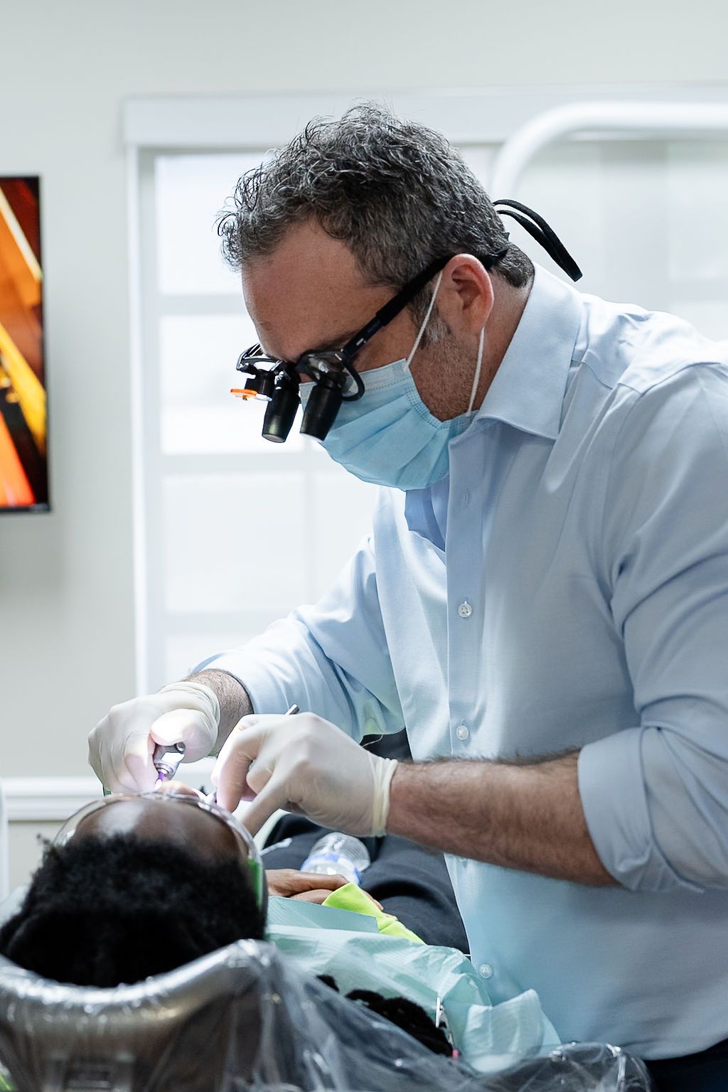 Dentist wearing magnifying loupes and mask treats a patient in a bright clinic room
