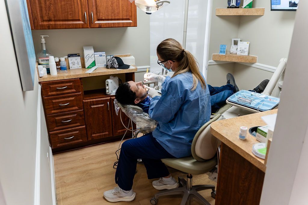 Dentist examining a patient in a clinic, with cabinets and dental tools visible.