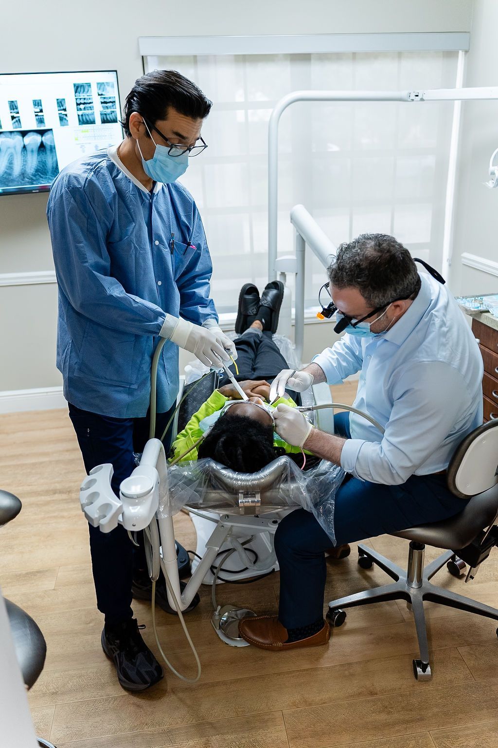 Two clinicians in blue PPE adjust a patient on a chair in a medical clinic.