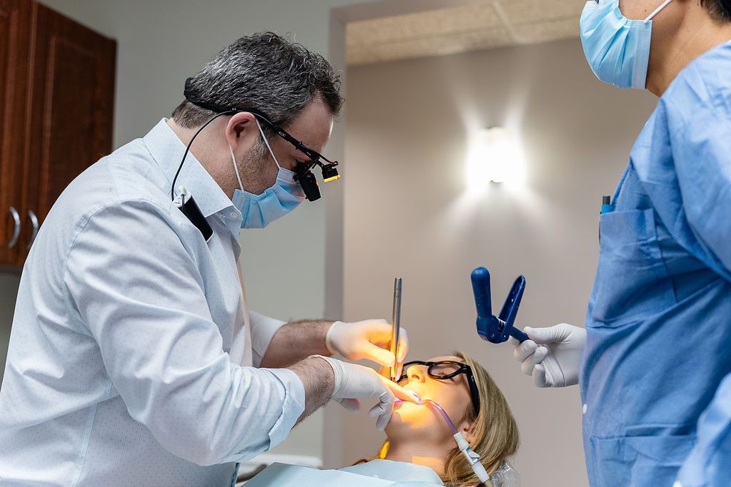 Dentist treating a patient in a clinic, wearing a mask and face shield while an assistant stands nearby