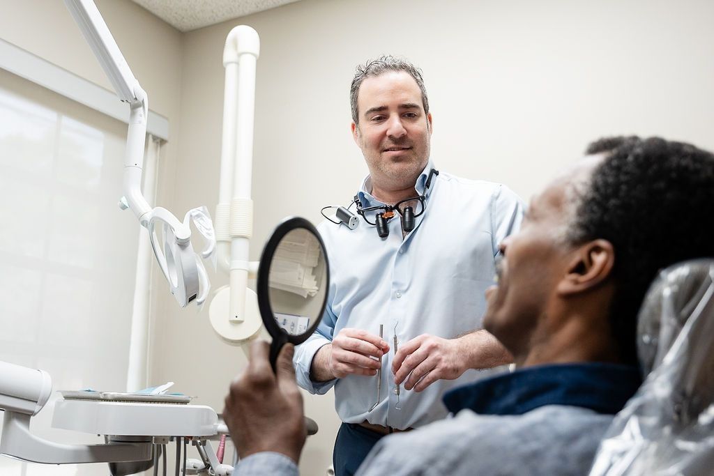 Dentist speaking to a seated patient in a dental clinic, holding a mirror near the exam chair.