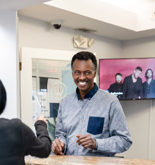 Smiling man in a blue shirt standing in a bright room, with others visible on a wall-mounted screen behind him