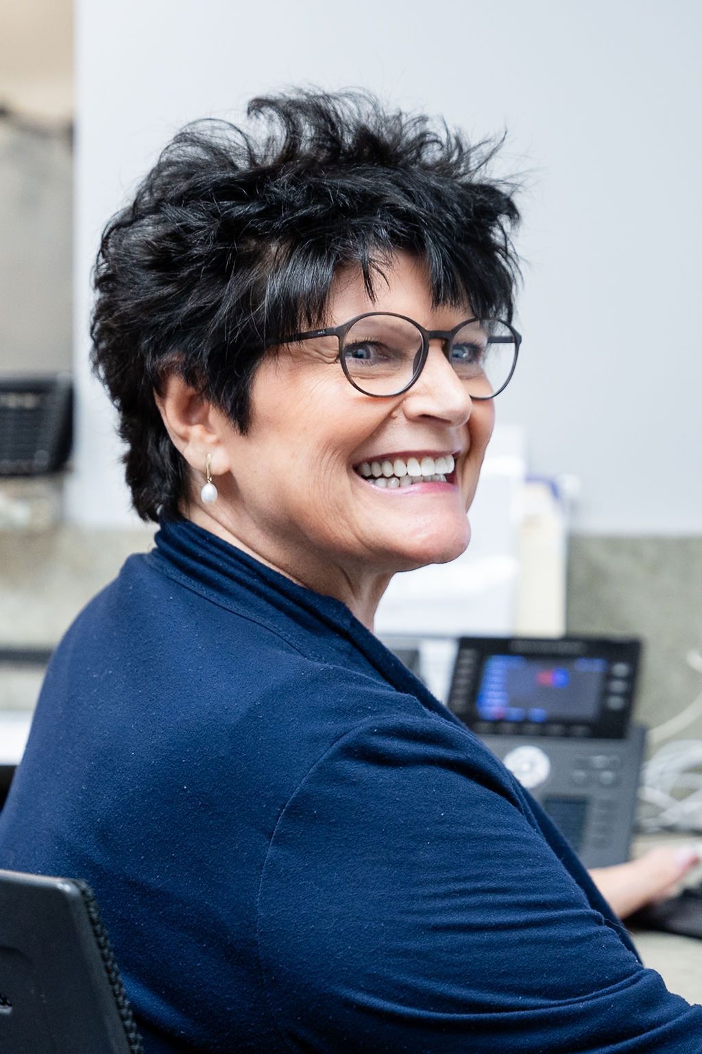 Smiling person with glasses seated at a desk in a navy sweater, looking over their shoulder in an office.