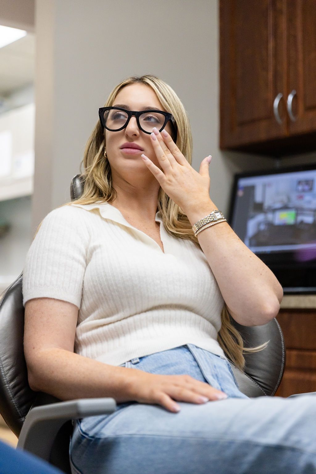 Woman seated in a dental chair, adjusting her glasses and looking thoughtful.
