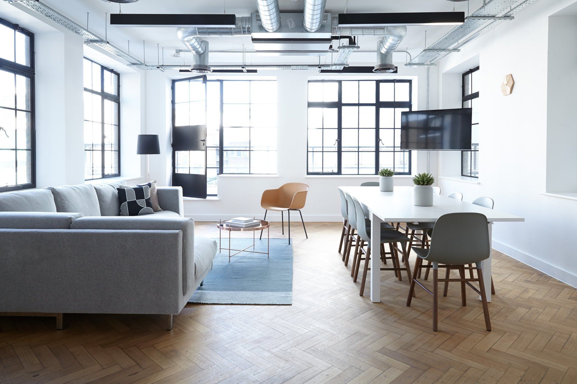 Bright loft-style living and dining room with gray sofa, wood table, large windows, and blue rug