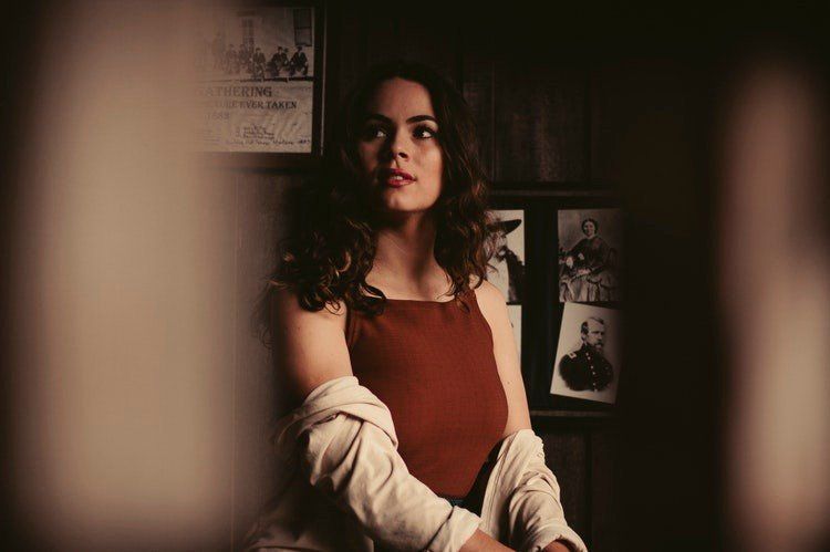 Woman in a red top with a light jacket, standing indoors against a dim wall with framed photos.