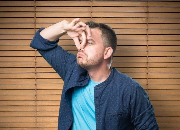 Man in blue shirt holding fingers to his nose, standing before wooden blinds.