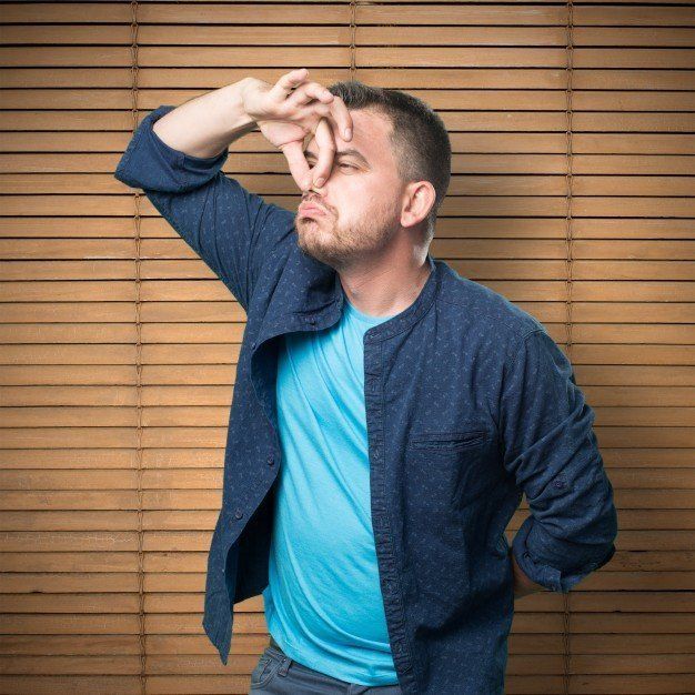 Man in blue shirt and jacket pinching his nose in front of wooden blinds