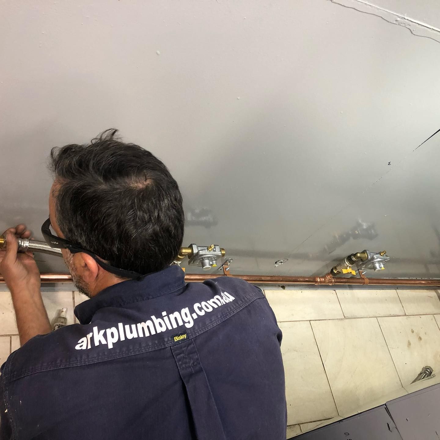 A Man Wearing A Blue Shirt With The Word Plumbing On The Back Is Working On A Pipe — ARK Plumbing & Gasfitting In Exeter, NSW