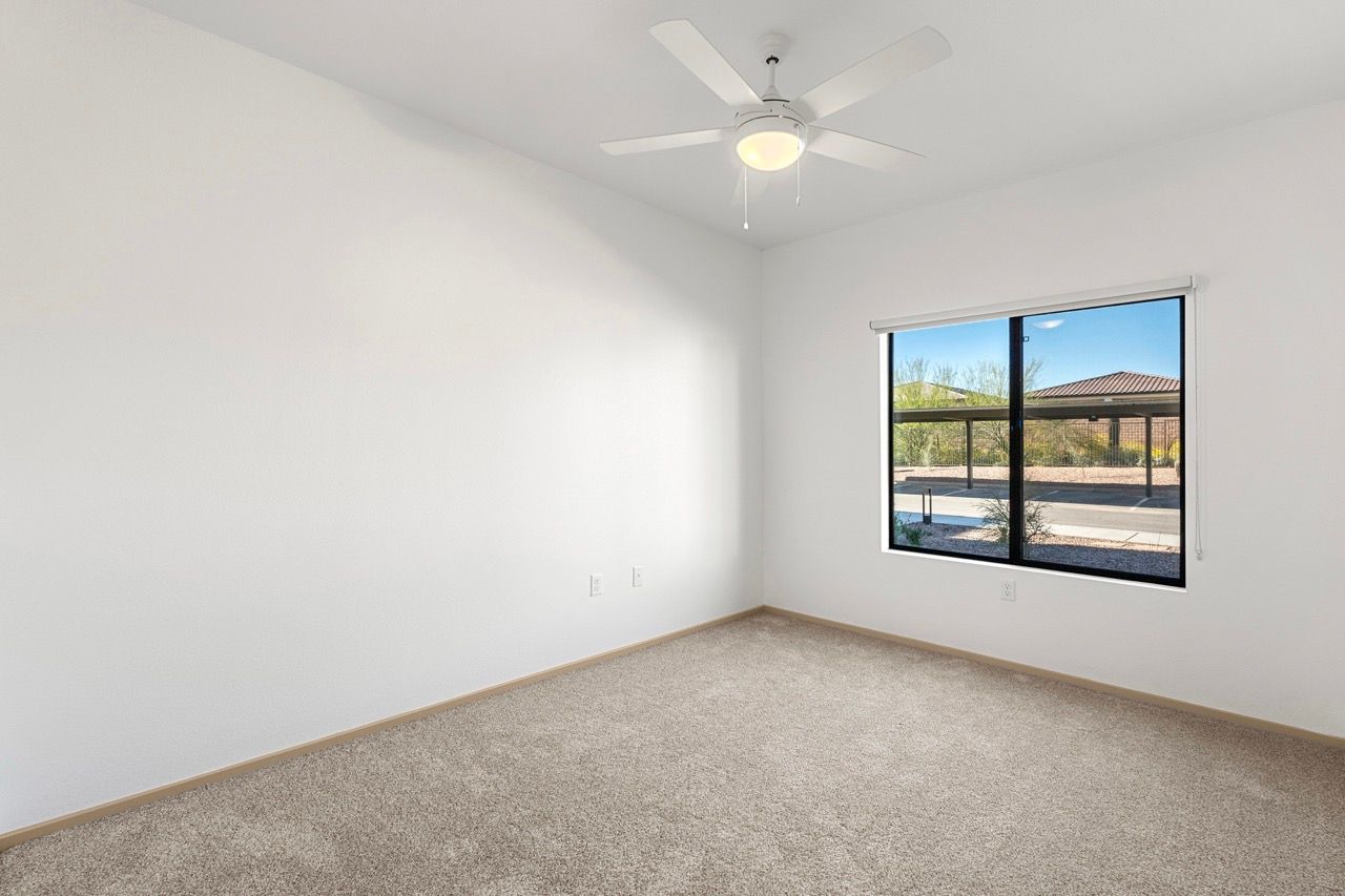 Empty bedroom in an apartment with a window, ceiling fan, and beige carpet.