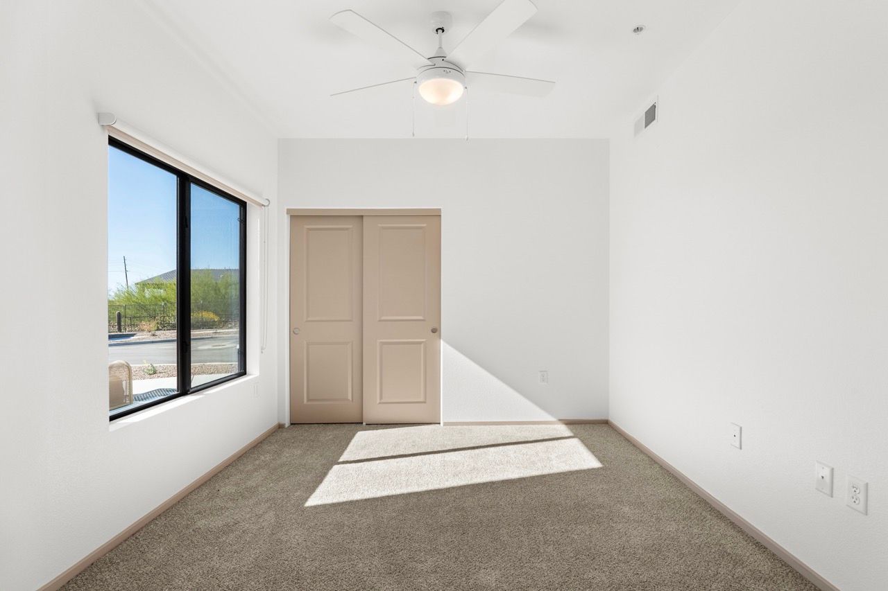 Empty bedroom with large window, beige double doors, ceiling fan, and neutral carpet.