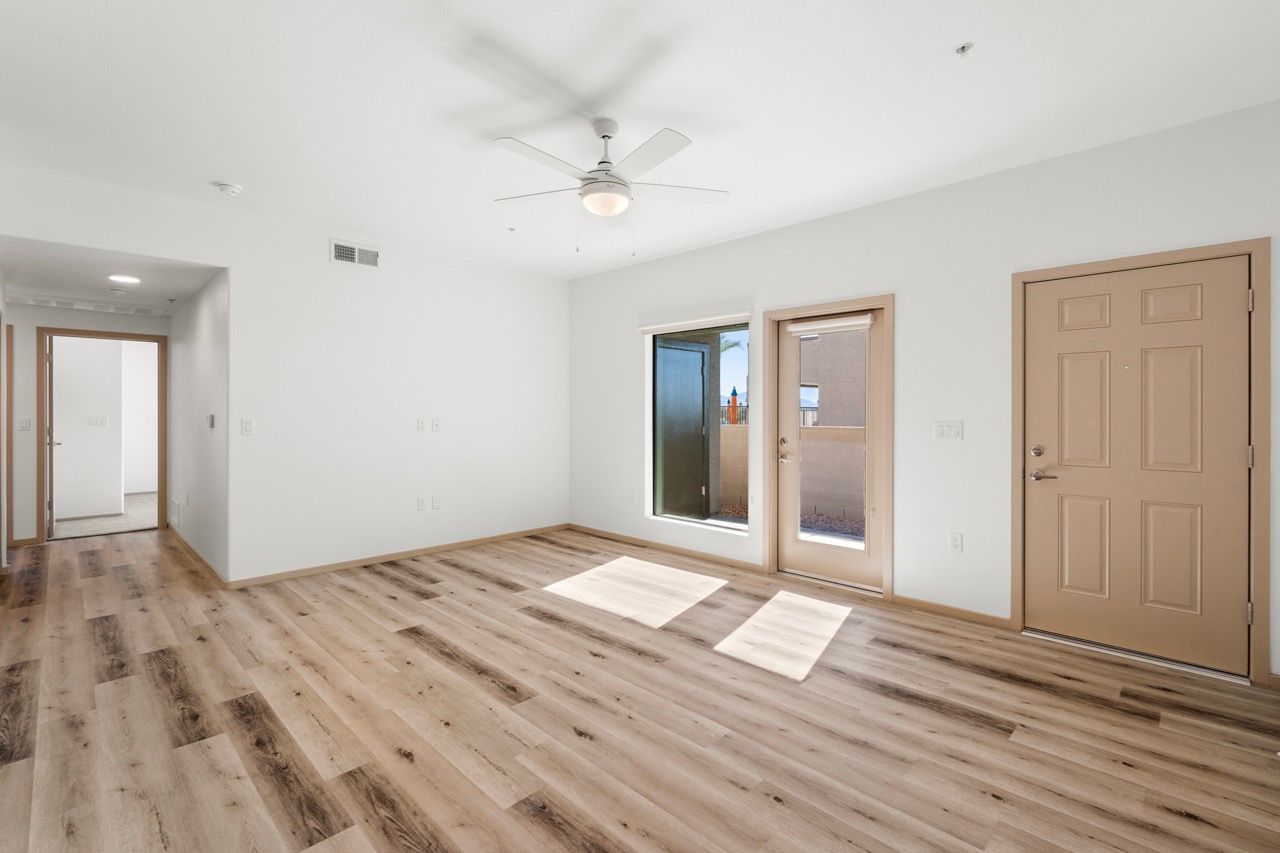 Empty living room with light wood flooring, white walls, ceiling fan, and exterior door.