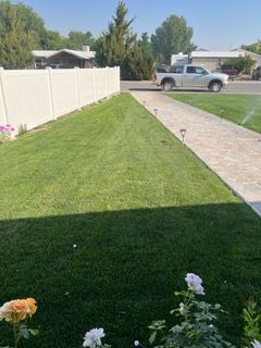 Green lawn beside a white fence and concrete path, with flowers in the foreground and a parked truck across the street