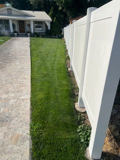 Narrow grassy strip beside a white fence, next to a stone driveway leading to a house.