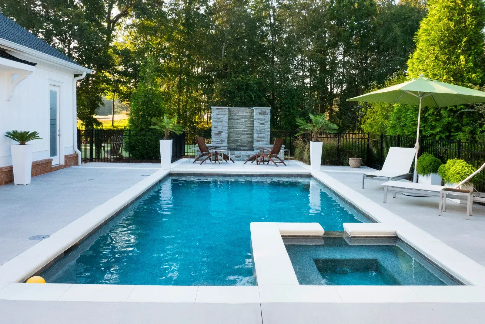 Modern backyard pool with lounge chairs, green umbrella, and hot tub beside a white house