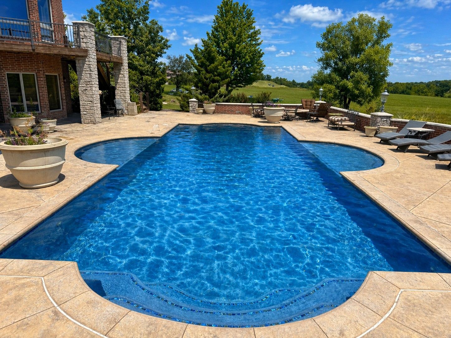 Backyard pool with blue water, stone patio, lounge chairs, and landscaped greenery under a clear sky