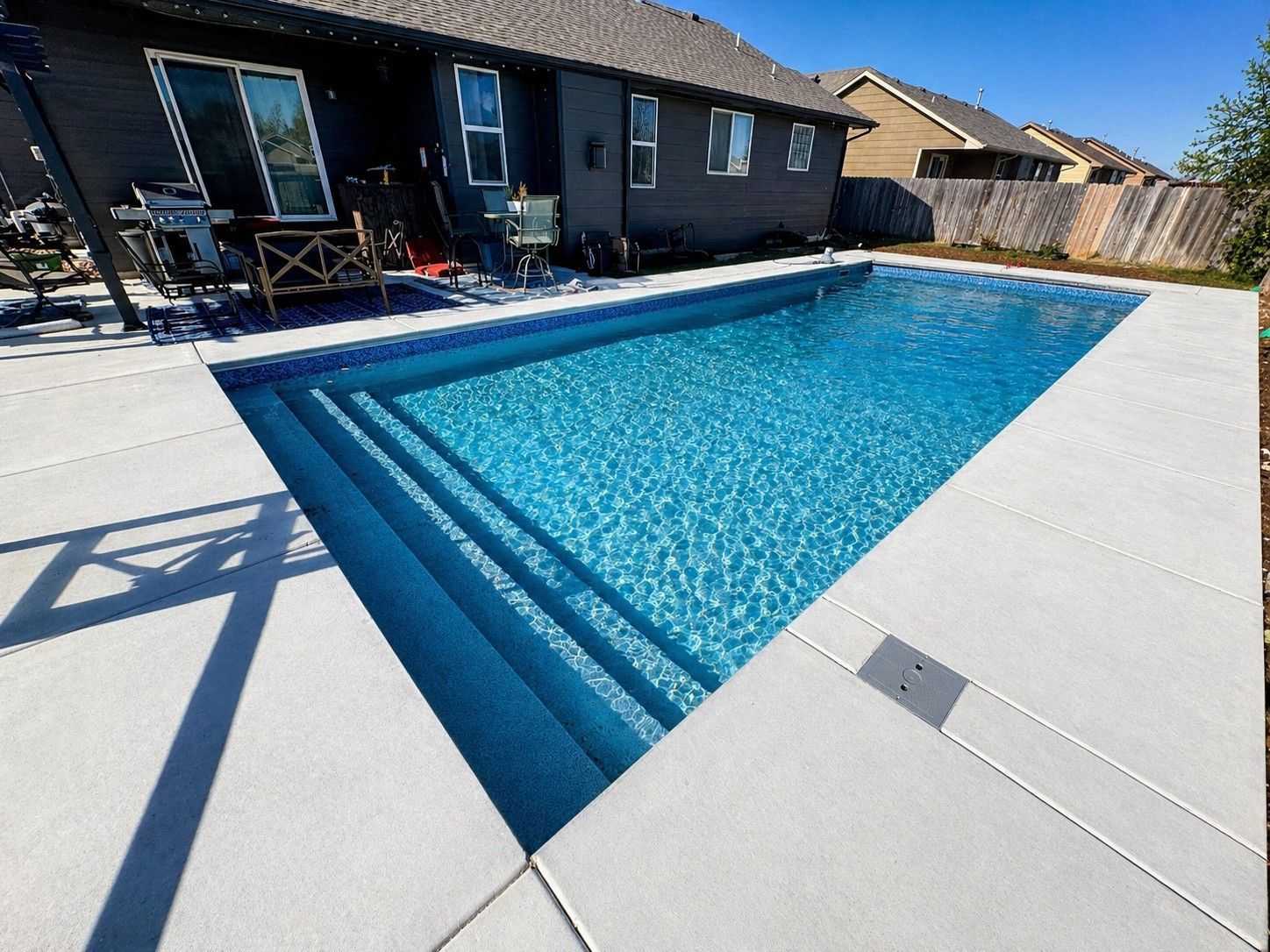 Backyard swimming pool with blue water and white concrete deck beside a house