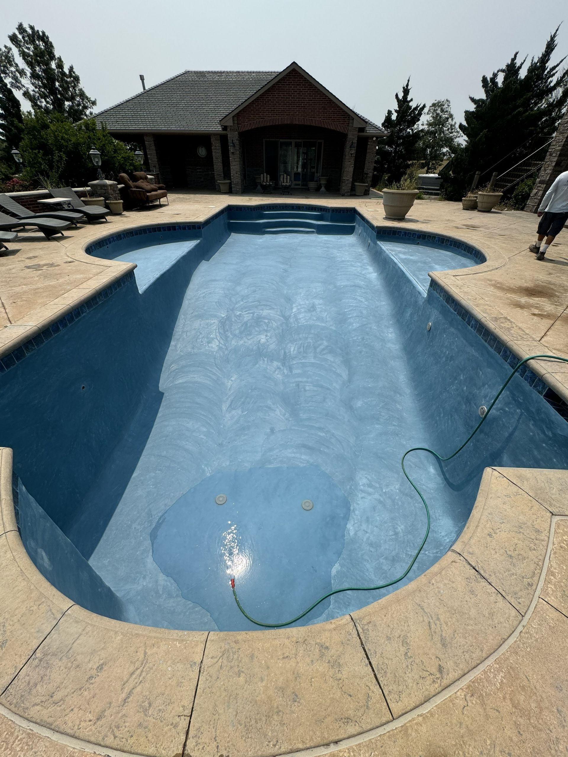 Empty blue swimming pool with a tan stone deck and a house in the background