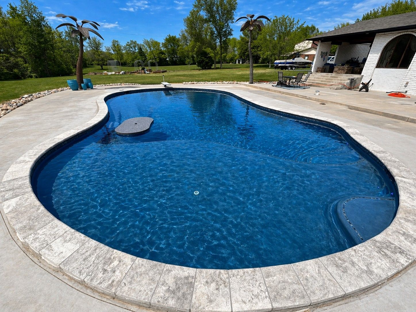 Curved backyard swimming pool with blue water, stone deck, and patio area under a clear sky