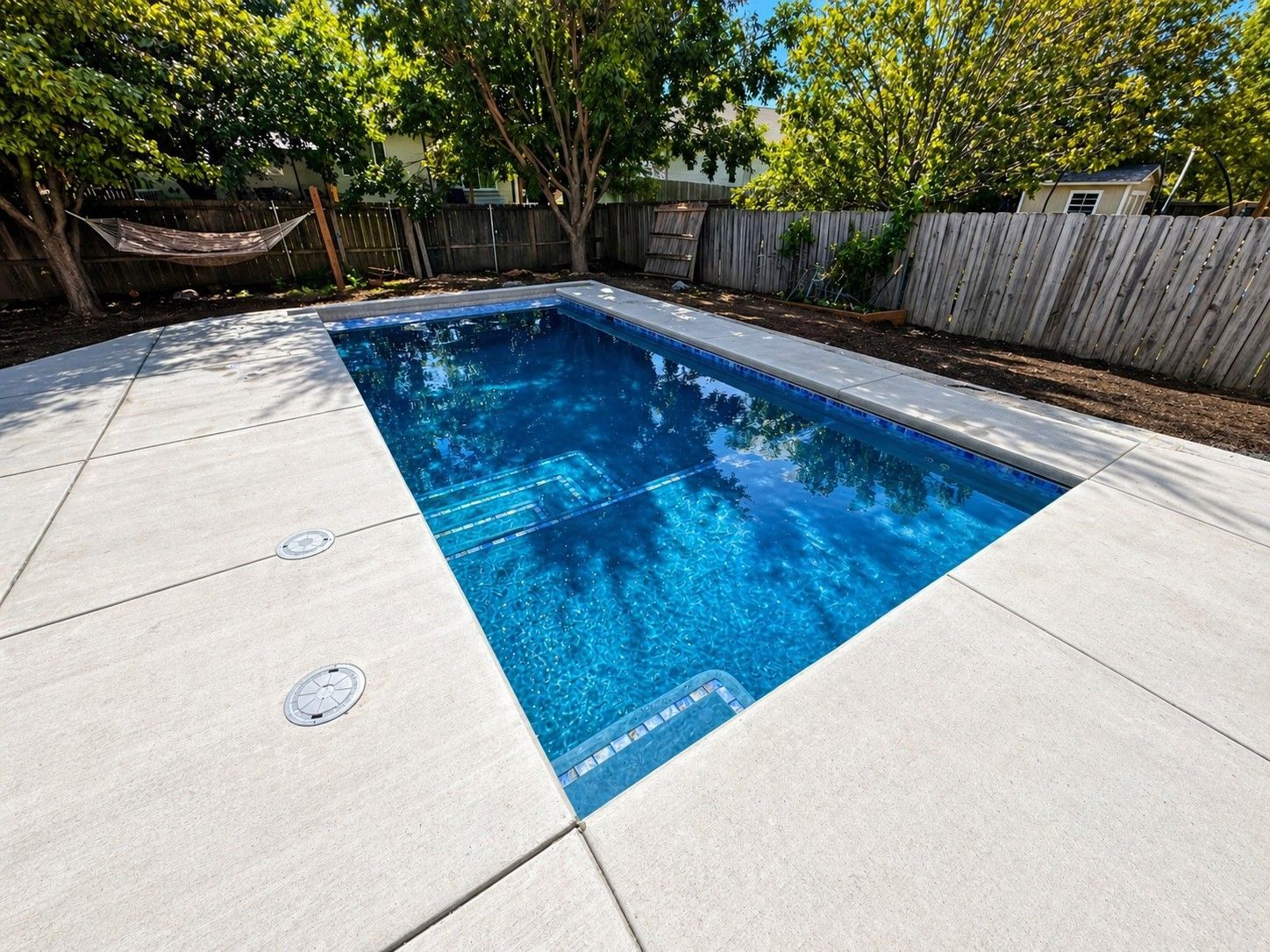 Sunny backyard pool surrounded by concrete, trees, and a fence.