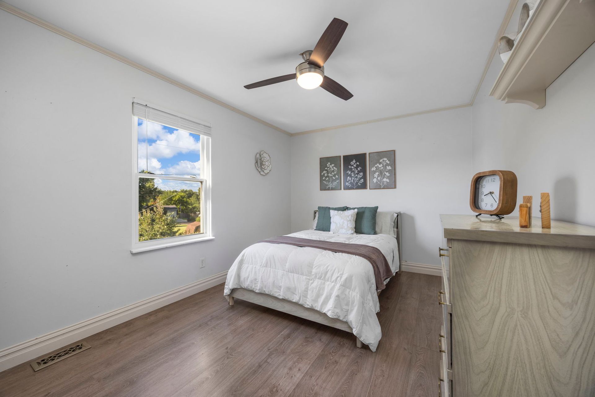 Bedroom with a bed, window, dresser, and ceiling fan. The walls are light gray and the floor is wood.