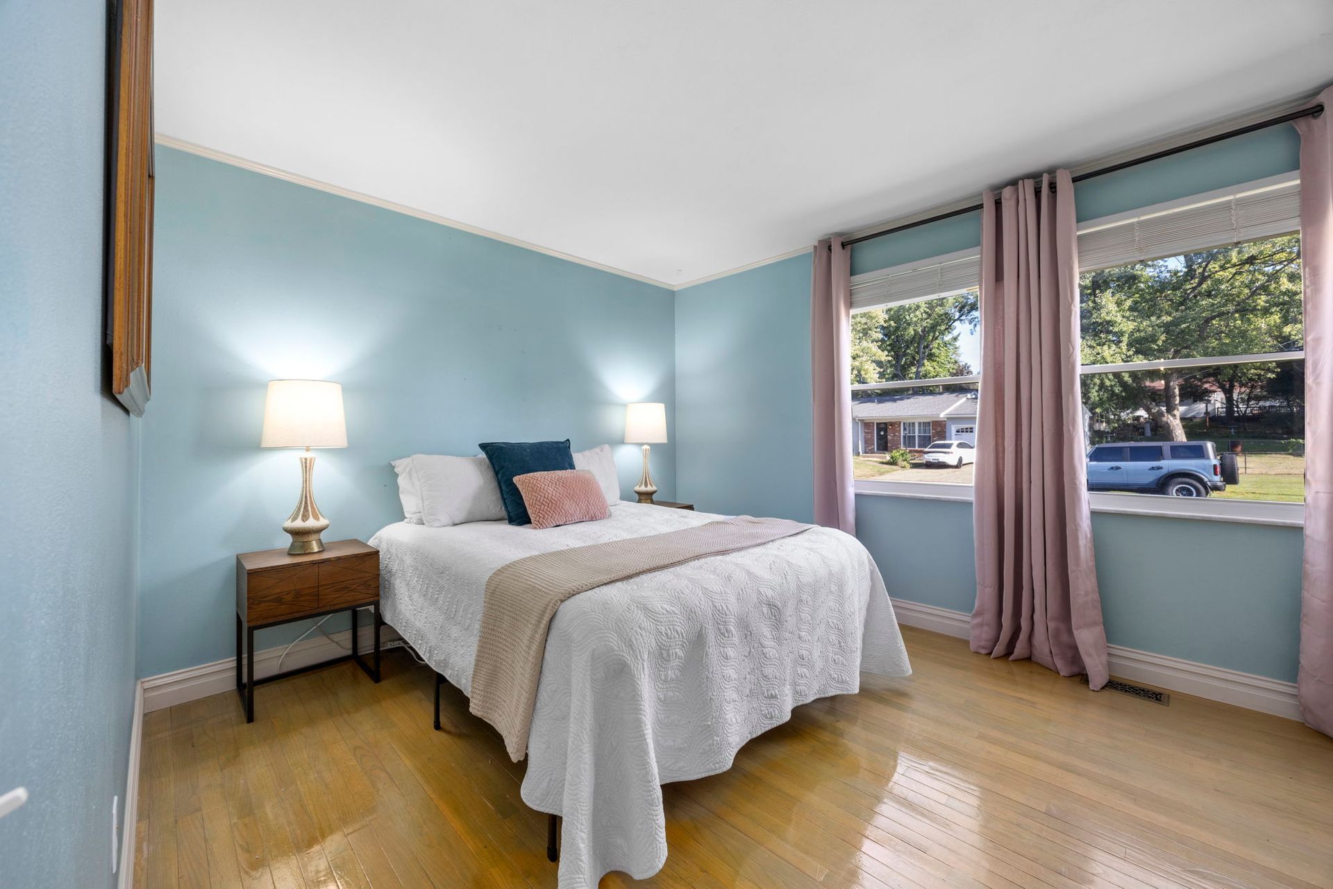 Bedroom with light blue walls, light wood floor, bed with white bedding, pink curtains.