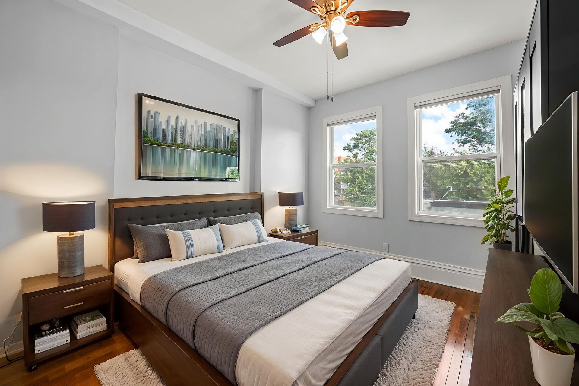 Bedroom with a wooden bed, two windows, and a TV. Light blue walls and a ceiling fan.