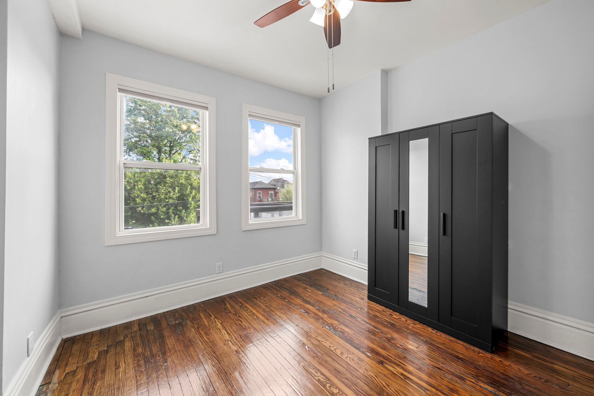 Empty bedroom with hardwood floor, two windows, and a black wardrobe with a mirror.