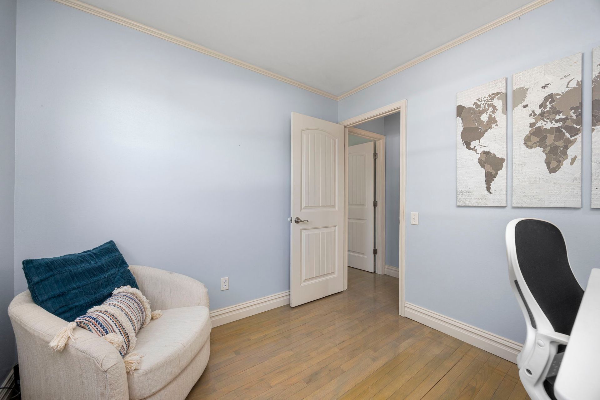 Small, blue-walled office with a chair, desk, world map art, and an open door leading to another room.